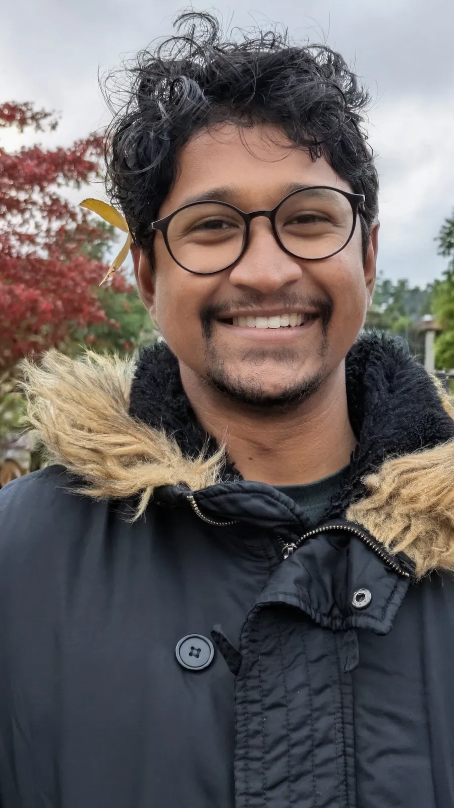 Sadat Daniel smiling outdoors in a black parka, autumn trees behind